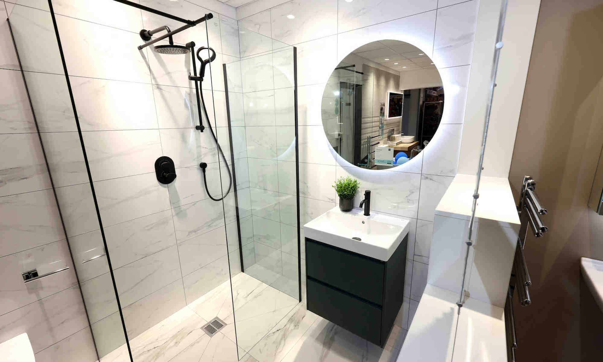 A modern style bathroom featuring pale marble tiles, a walk in shower, sink on top of a black cabinet and a round mirror on the wall